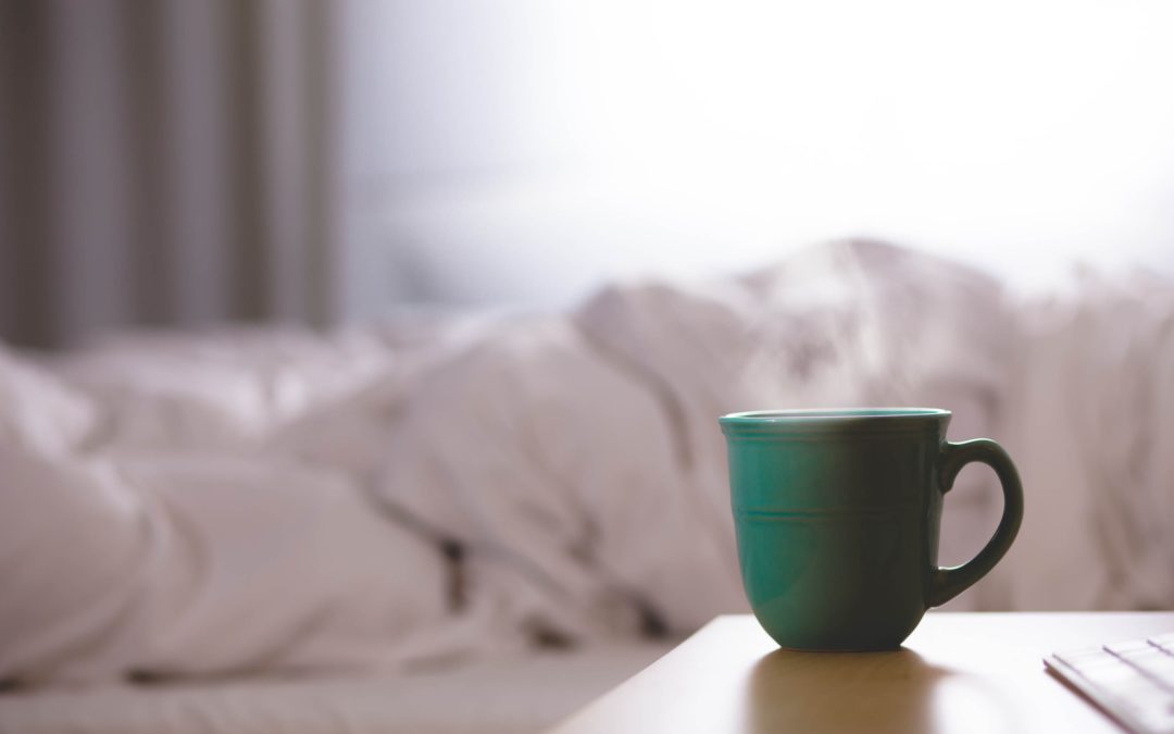 Image of a green coffee mug on a bedside table and a white, rumpled duvet on the bed in the background