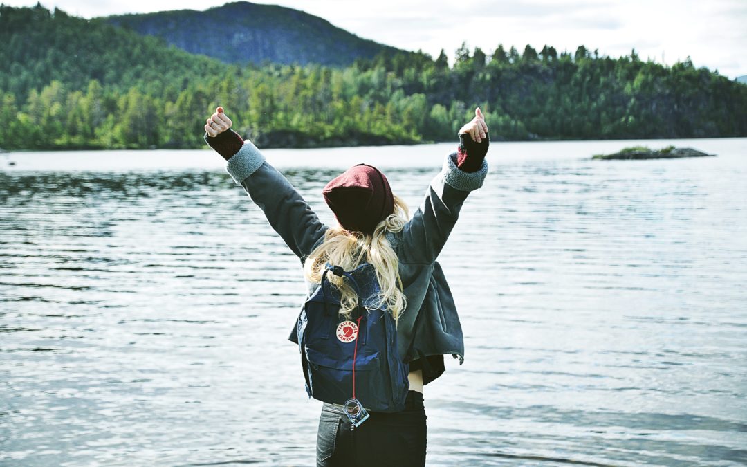 A woman wearing a toque and a backpack with both arms in the air facing towards a lake with mountains in the background