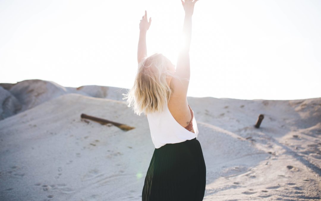 Image of a blonde woman standing facing away from the camera at 45 degrees with both hands in air on a sunny day