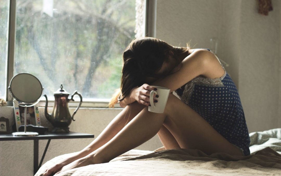 Image of a woman with brown hair sittlng on her bed holding a coffee mug with her heads on her knees looking sad