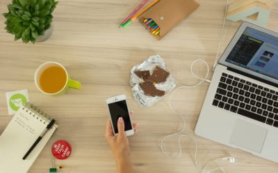 Image of a woman's hand holding a cell phone over a desk filled with many items, including a laptop, chocolate bar, notepad and pen, coffee, plant and other office supplies