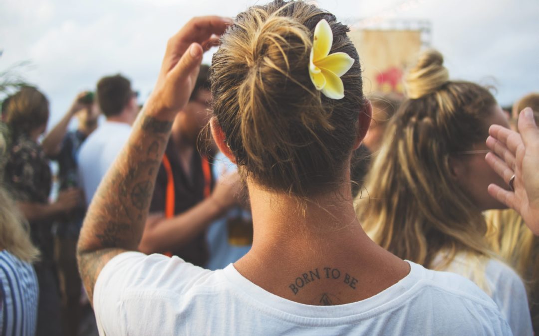Photo of the back of a woman's head with blond hair up in a bun with a yellow flower on the side scratching her head with a number of other people in front of her also facing away from the camera