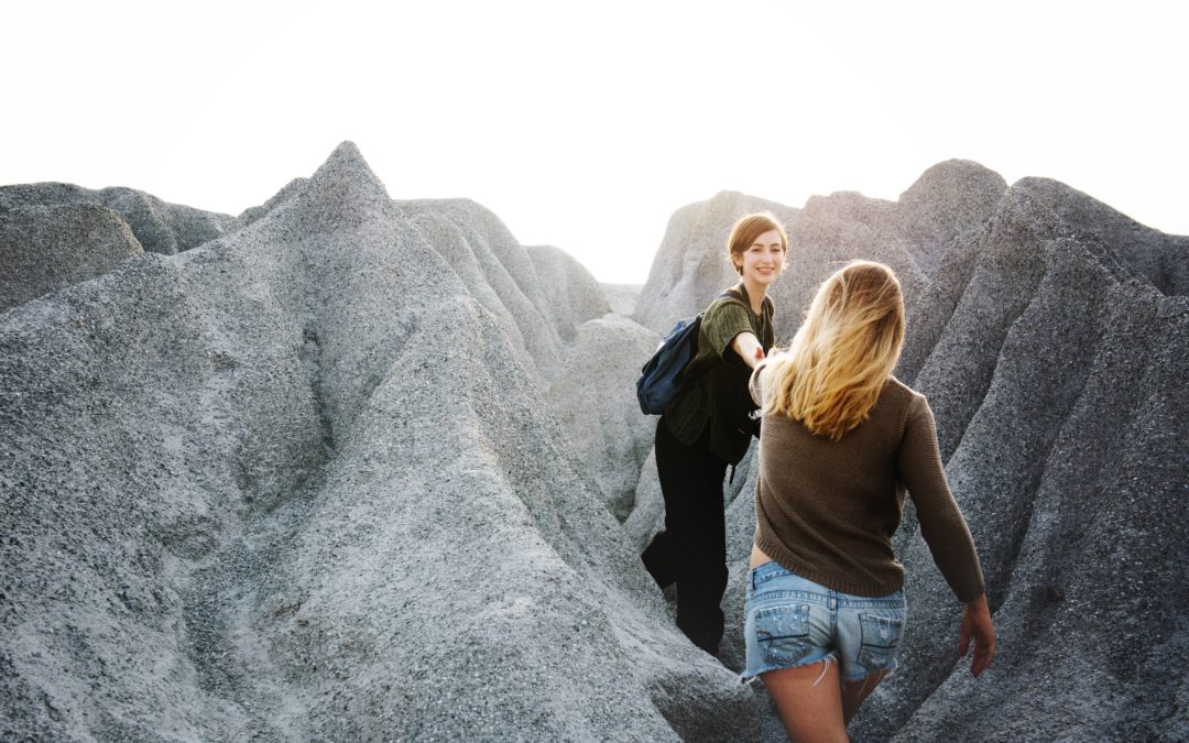 Photo of two women hiking in rocky mountains with one woman turning around and offering a helping hand to the other woman