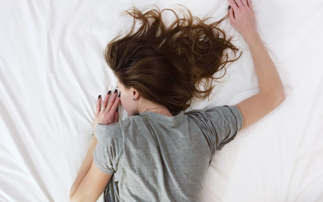 Overhead photo of a woman wearing a grey t-shirt and long brown sleeping stomach down with her head to the left side on a white sheet