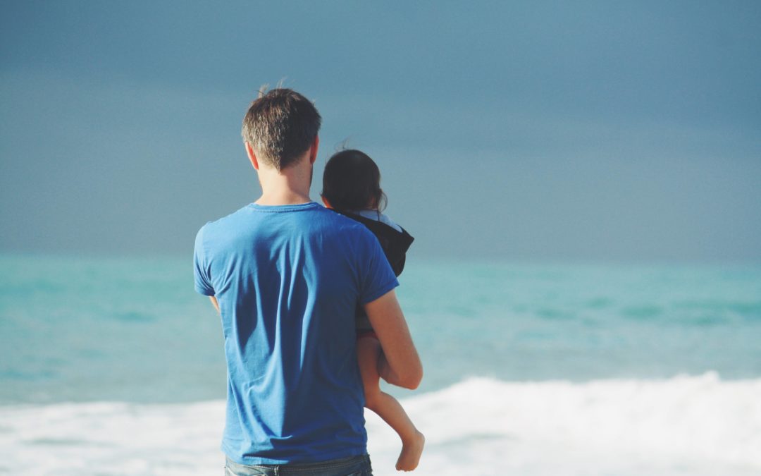 Image from behind of a father holding his infant son as they look out towards the ocean on a sandy beach