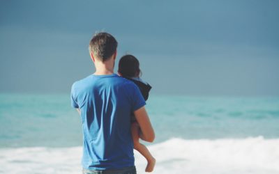 Image from behind of a father holding his infant son as they look out towards the ocean on a sandy beach