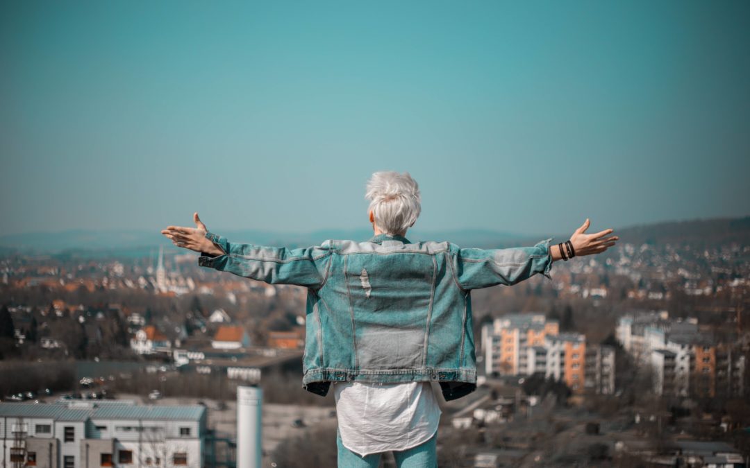 Photo of the back of a man with white hair with arms outstretched on a hill looking out over a city