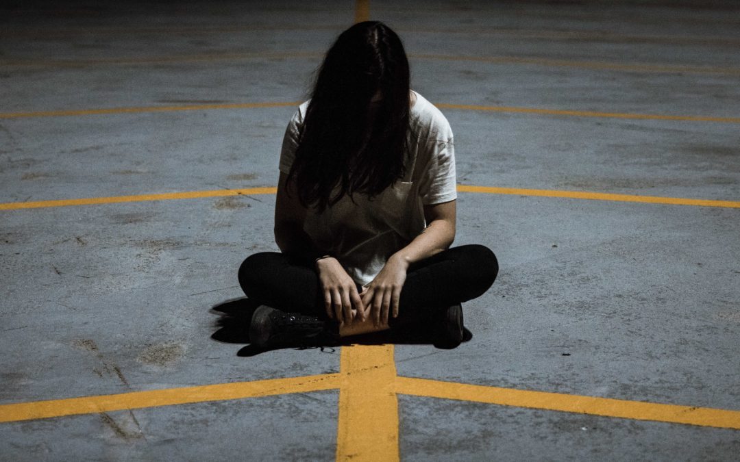 Photo of a woman sitting cross legged in a parking lot on cement with yellow parking lines with her head hanging down so her hair is covering her face