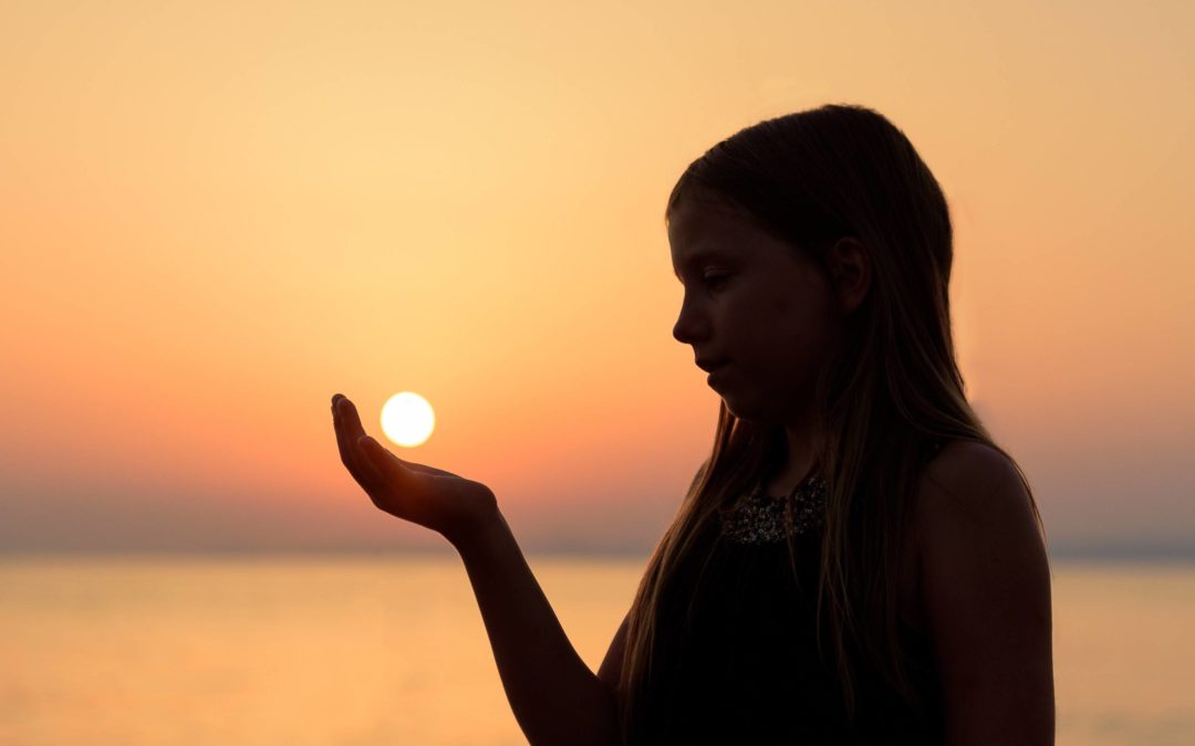 Image of the silhouette of a young girl with long hair cupping her hand out in front of her with the sunset in the background so it looks like she is holding the sun in her hand