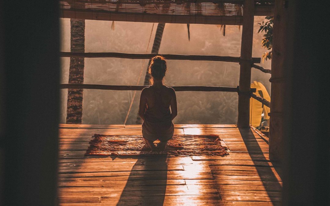 Photo taken from behind of a woman meditating in the early morning sun on a wooden floor looking out at a forest
