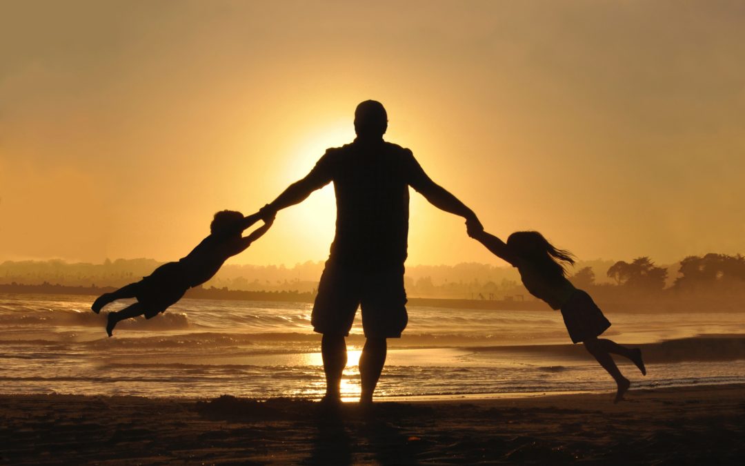 Silhouette of a father swinging one child in each hand around against a sunset on a beach