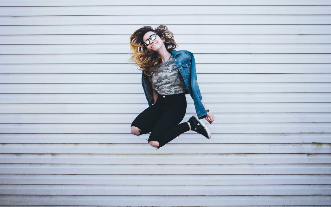 Photo of a smiling young woman wearing black ripped jeans, a white shirt and a blue jean jacket who is jumping in the air with her knees bent and arms down against a white horizontally striped background