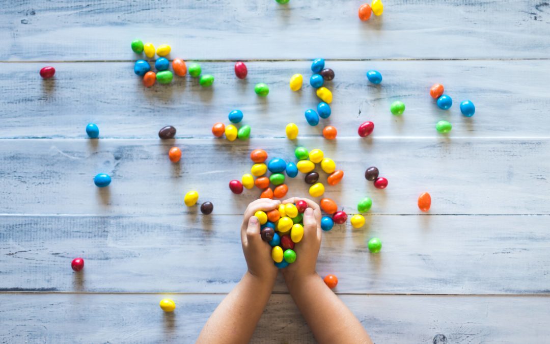 Overhead photo of a child's hands holding peanut M & Ms of different colours with some scattered onto the white table top