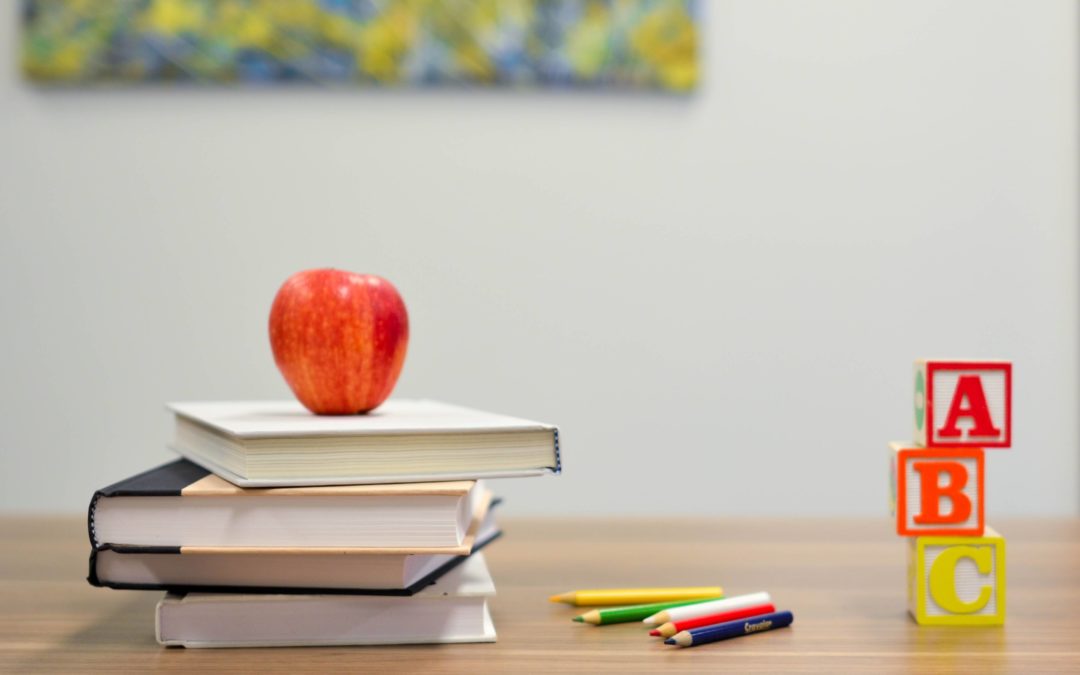 Image of a desk with pencil crayons beside a pile of books with a red apple on top