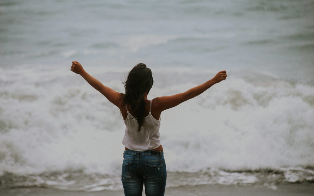 A woman with long brown hair wearing a white tank top and jeans stands looking out at the ocean with her two arms outstretched above her head