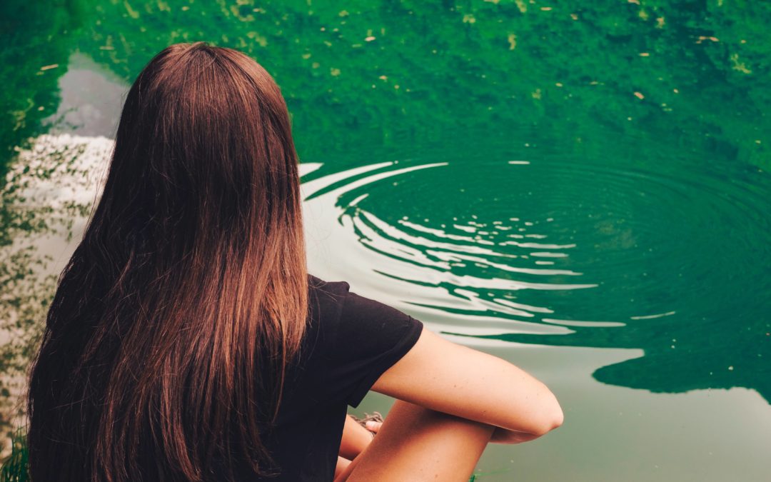 A woman with long brown hair sitting cross-legged and looking out at a green lake with a ripple on the top of the water