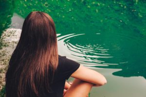 A woman with long brown hair sitting cross-legged and looking out at a green lake with a ripple on the top of the water