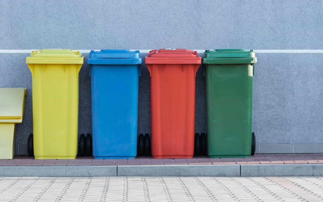 Four colourful outdoor garbage cans in yellow, blue, red and green in a row in front of a grey wall with a boardwalk in front