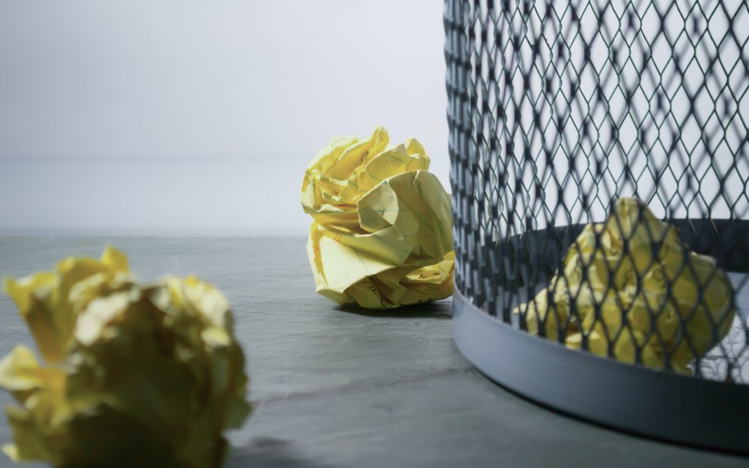 Grey metal trash bin with two crumpled up yellow sheets of paper on the floor beside the trash can and one inside the trash can to match theme of trashing good ideas to spur innovation