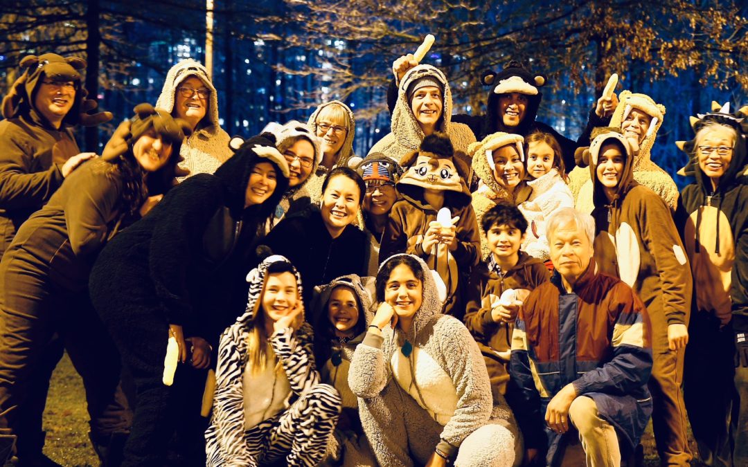 20 people in fuzzy animal pyjamas along with one man dressed in regular clothes against a backdrop of holiday lights