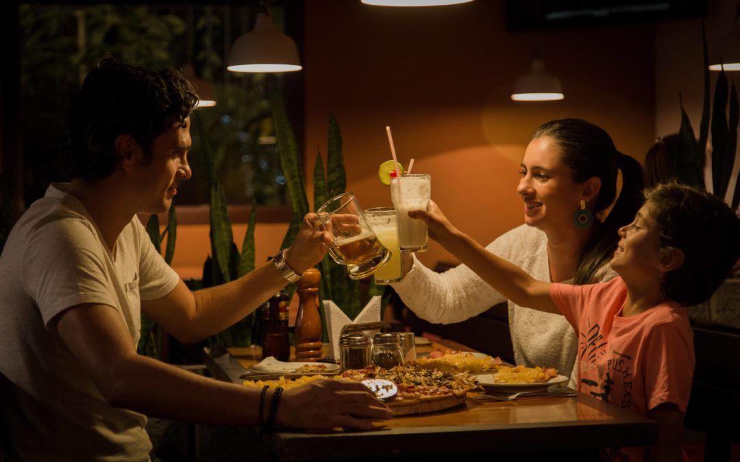 A father, a mother and child at a restaurant booth each holding their glasses up to clink them as a toast while smiling and laughing