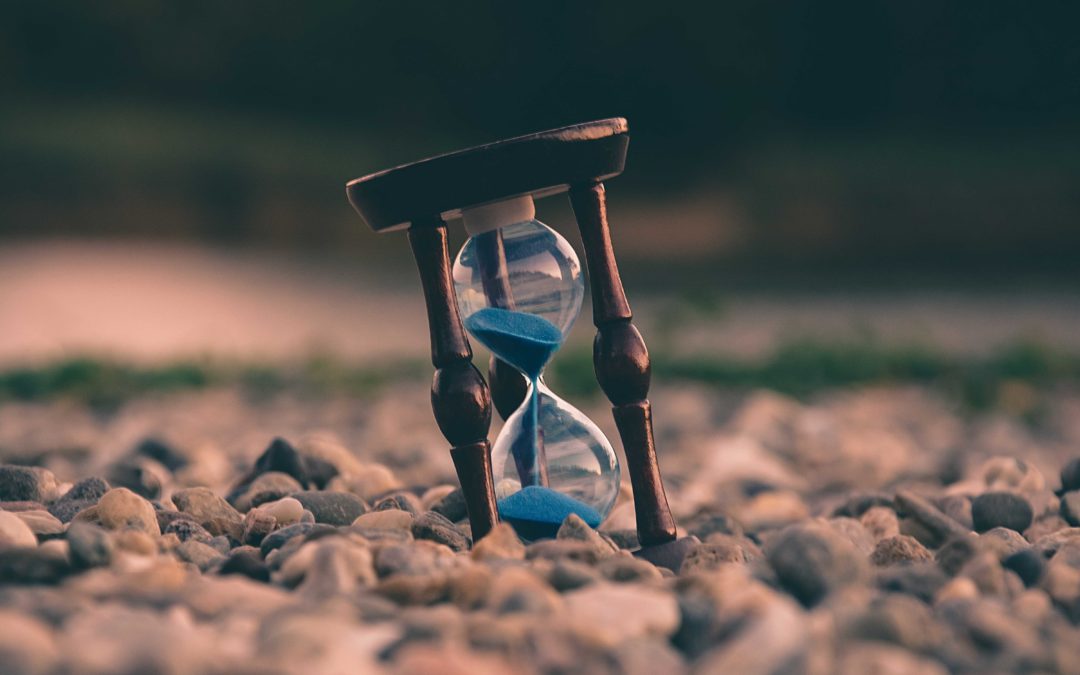 A wooden hourglass with blue sand inside sitting crookedly on a pebble beach