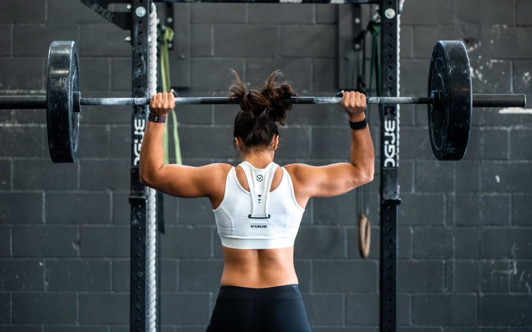 Back of woman in black tights and white top performing a overhead lift with a barbell with her biceps, shoulders and back muscles flexed