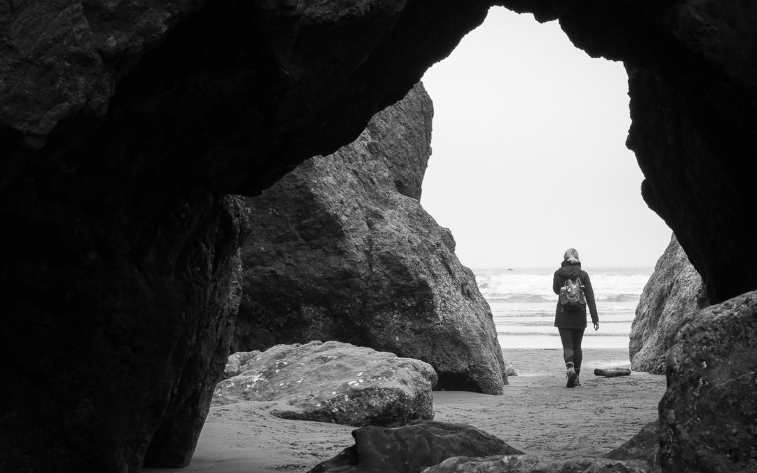 Black and White image of woman hiking through a rock cave and emerging onto a beach