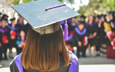 Photo from behind of woman with brown hair wearing a cap and gown with the audience facing her in the background