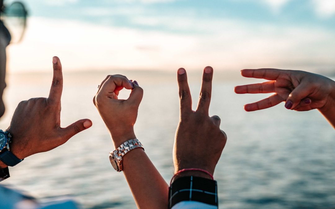 Two people holding up their hands in shapes that spell the word love against a background of a blue sky and beach