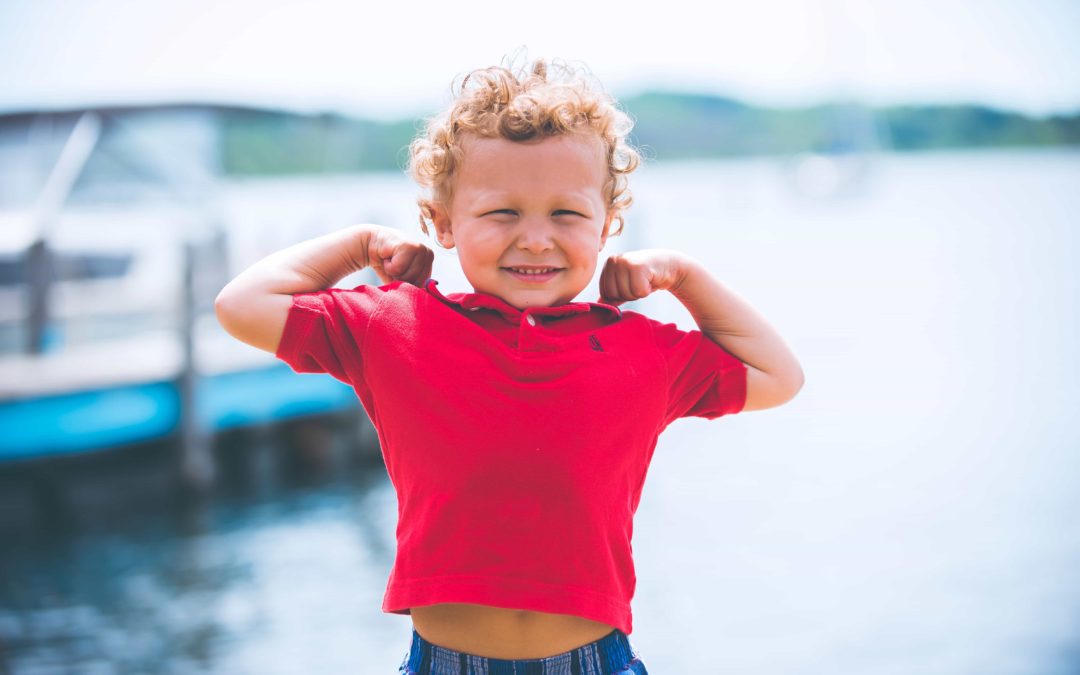 Little blond boy standing in front of a dock flexing both arms and smiling