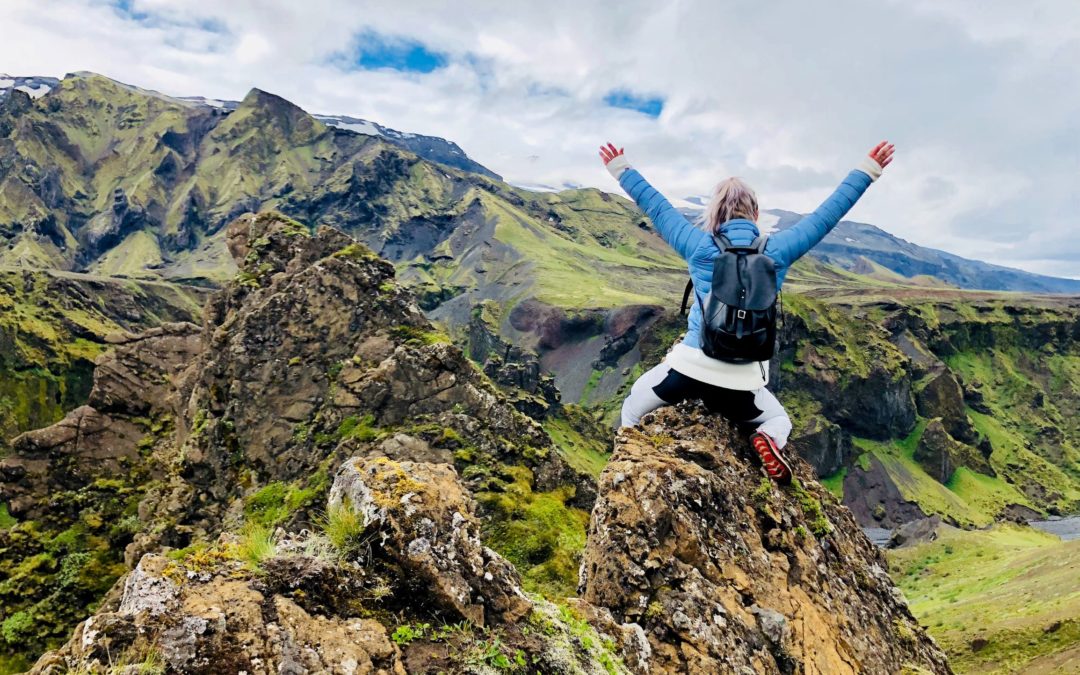 Woman sitting on top of a green mountaintop with both hands in the air victorious