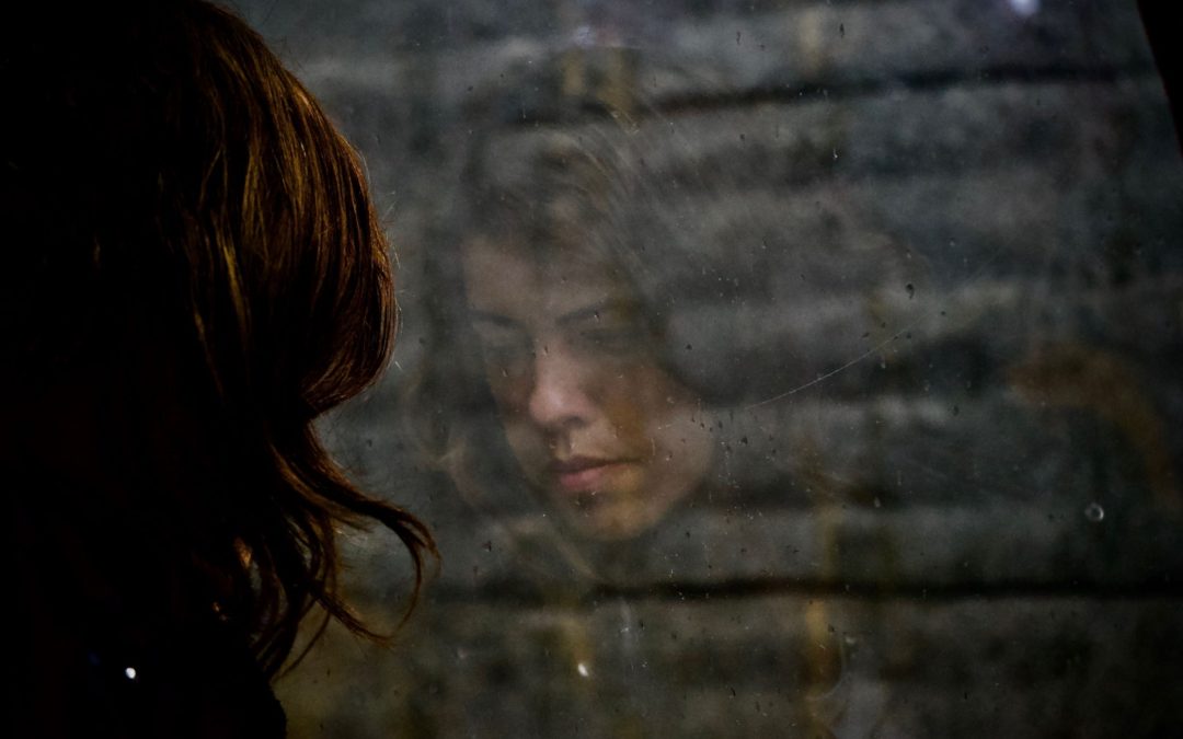 A woman with brown hair looking down with her reflection visible in a window looking out towards a grey brick wall