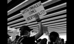 White man wearing a face mask holding up a sign at a protest that says: "With privilege comes responsibility"