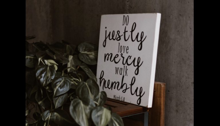 A canvas with the words "Do justly, love mercy, walk humbly" written on it in cursive writing on a table beside a plant with dark green leaves