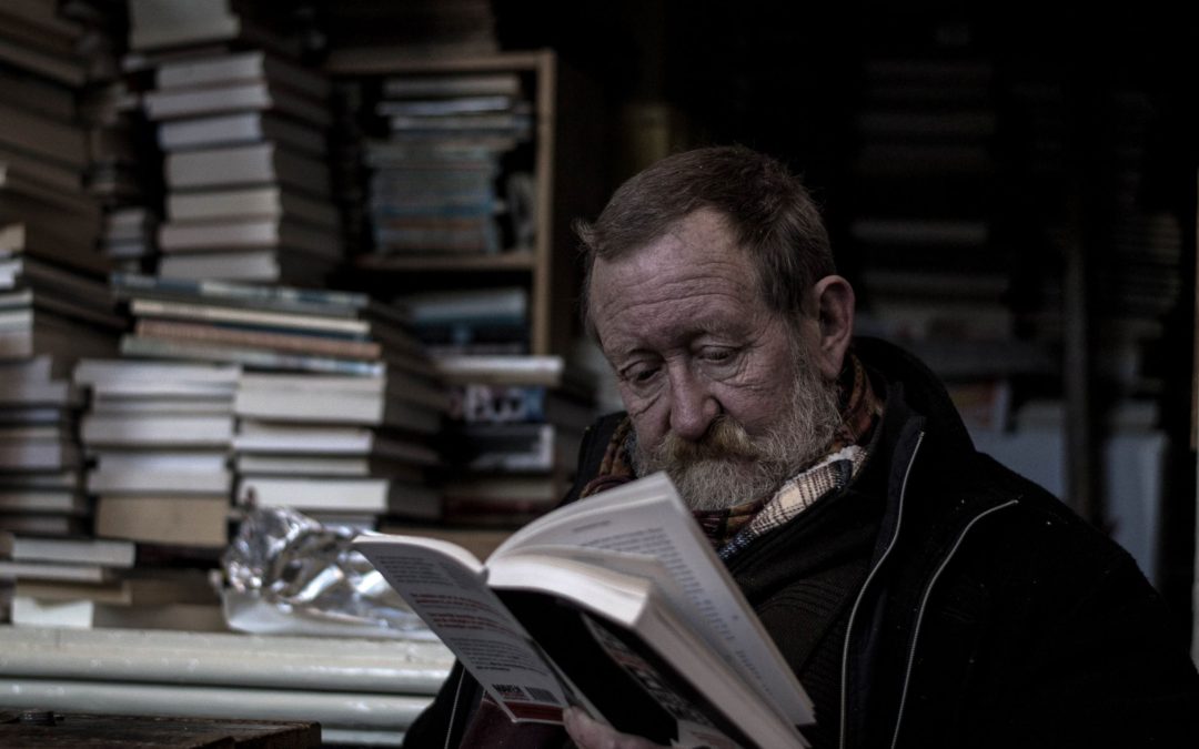 An older White man reading a book surrounded by piles of books