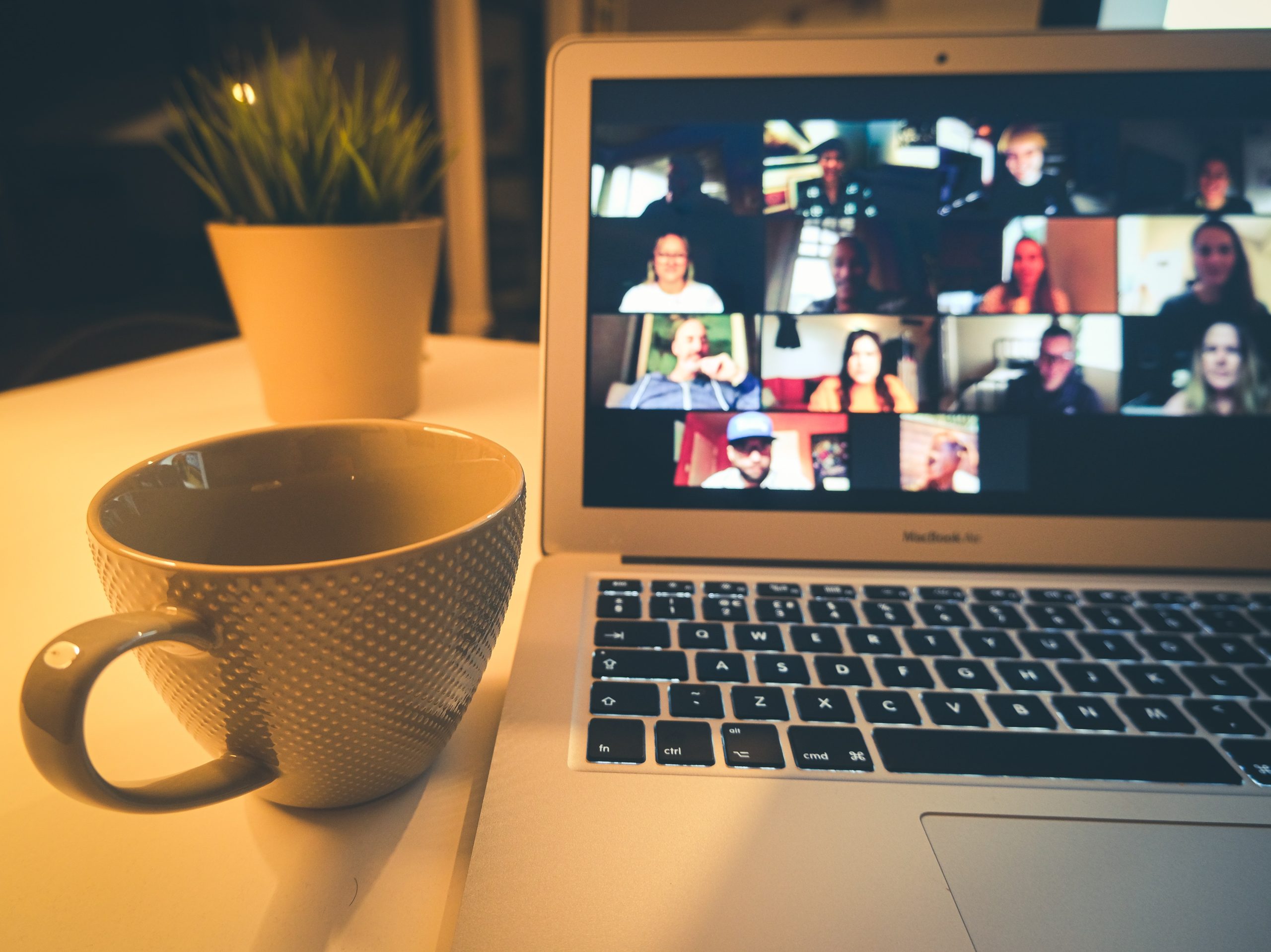 A desk with a mug, a plant and a computer with a screenshot of many zoom boxes