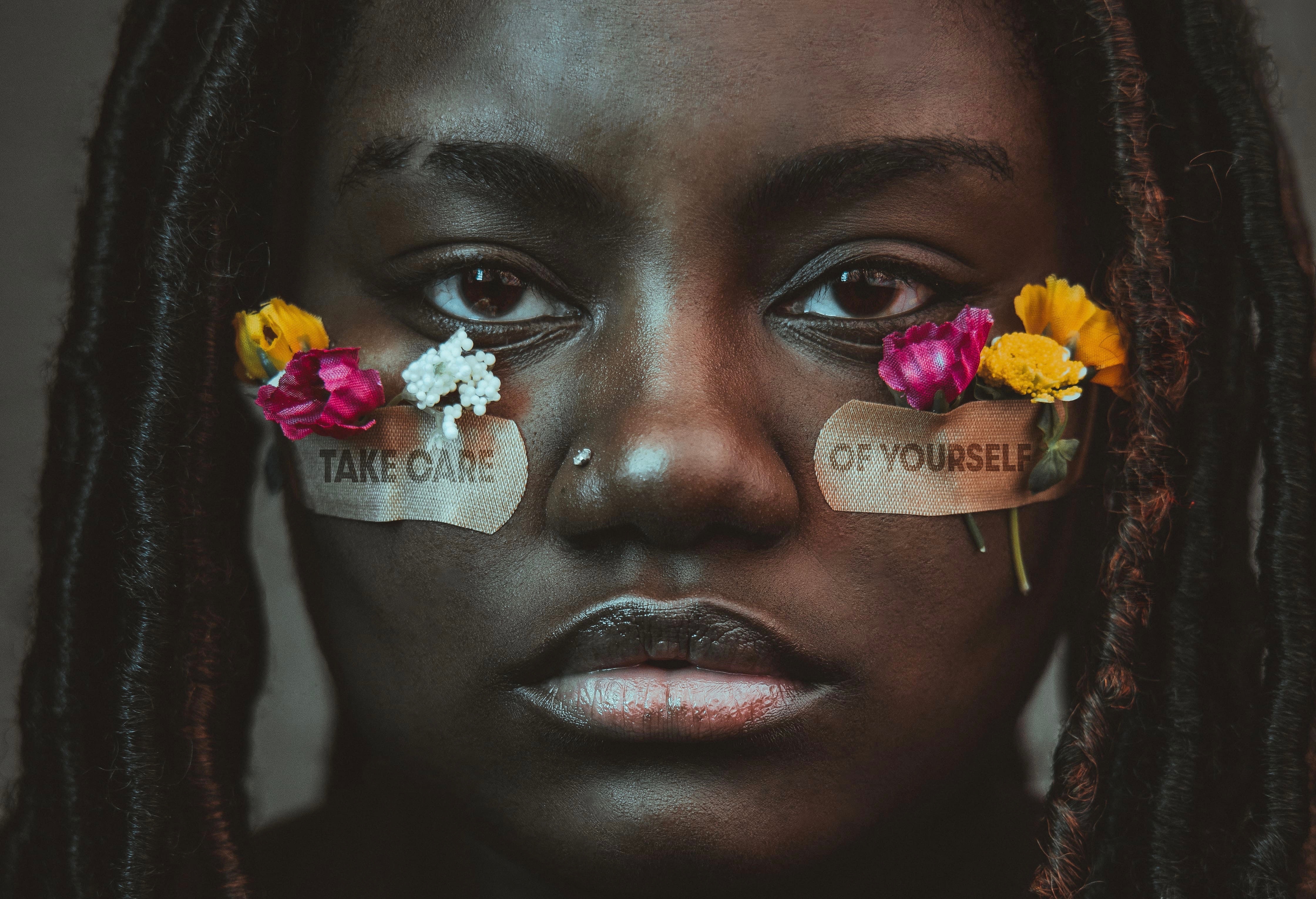 Close-up of a Black woman face with two bandaids holding flowers stuck under her eyes with the message "Take Care of Yourself" on the Bandaids