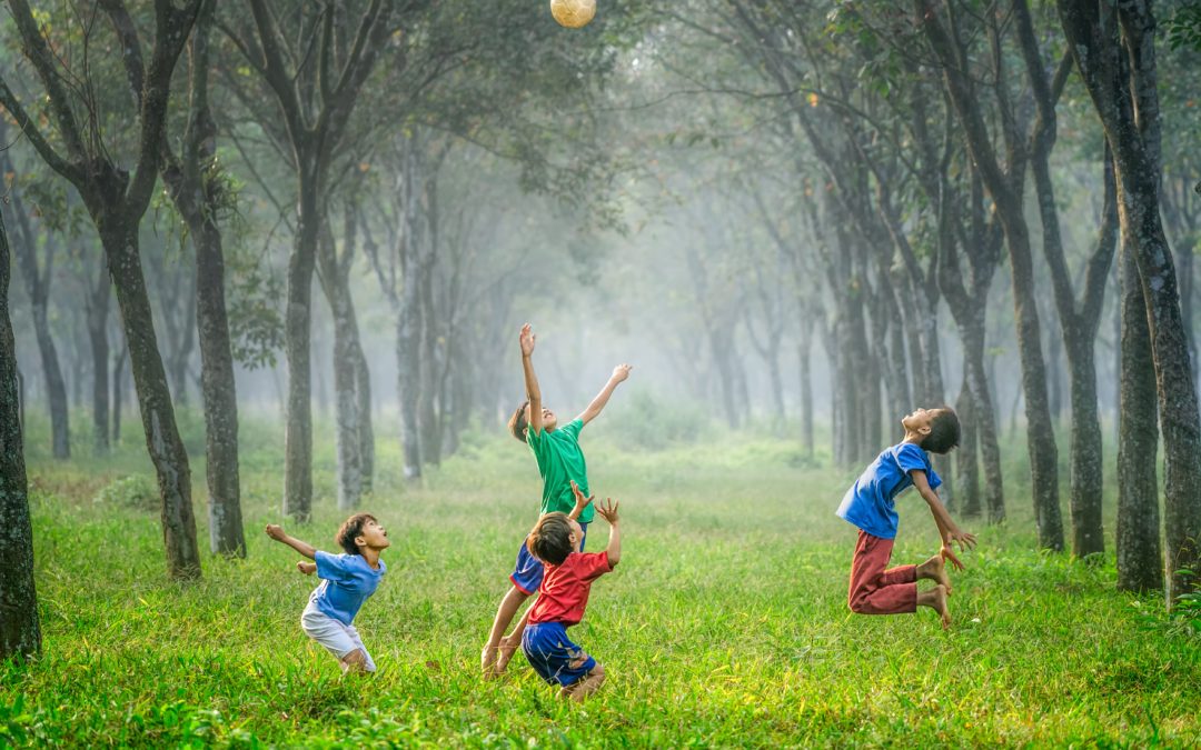 Four boys jumping up towards a ball high in the sky on grass surrounded with trees in the background