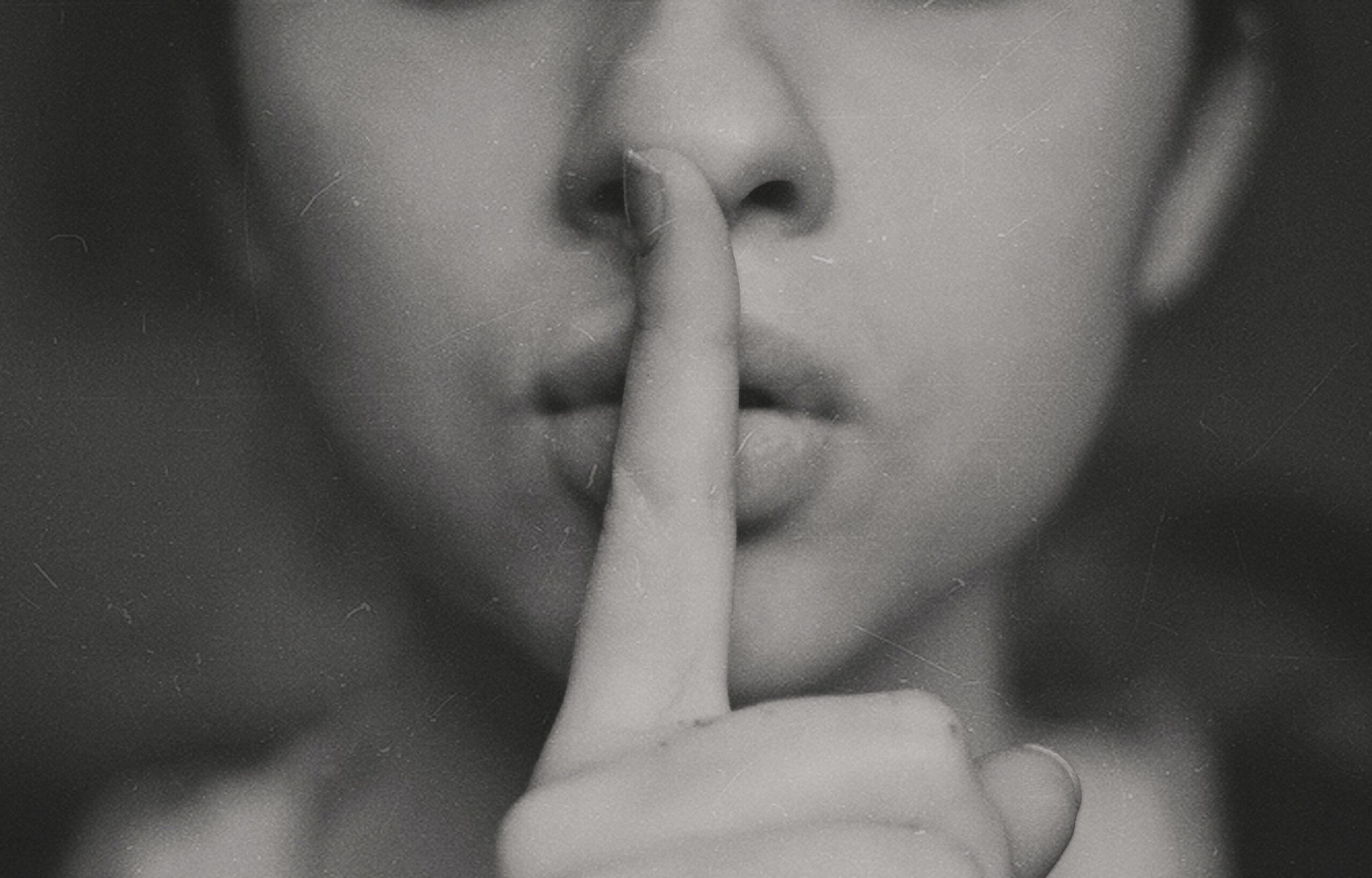 Photo of the bottom half of a woman's face with her holding her index finger in front of her lips indicating: "shhh"